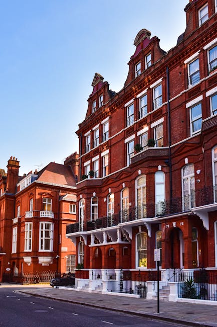 Exterior view of a red brick residential building with multiple stories, featuring large windows, some with decorative metal railings for balconies. The ground floor has white-framed doors with steps 