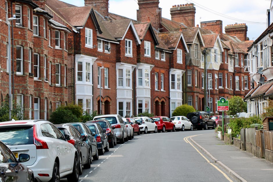Exterior view of a red brick residential building with multiple stories, featuring large windows, some with decorative metal railings for balconies. The ground floor has white-framed doors with steps 