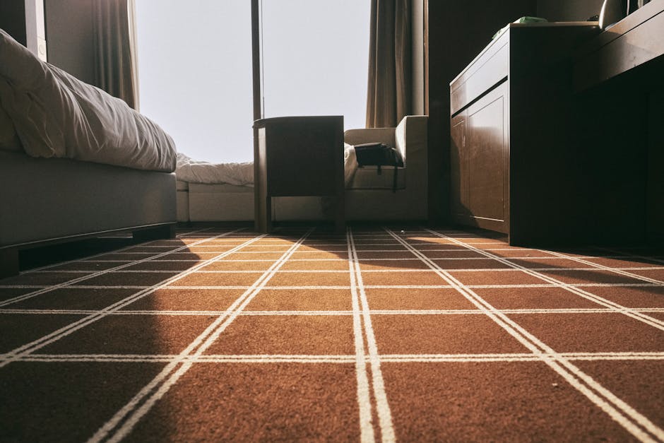 A close-up view of a bedroom carpet featuring a brown and beige checkered pattern, with visible linear grid design. The carpet covers a hardwood floor surface in front of a bed with white bedding, sit